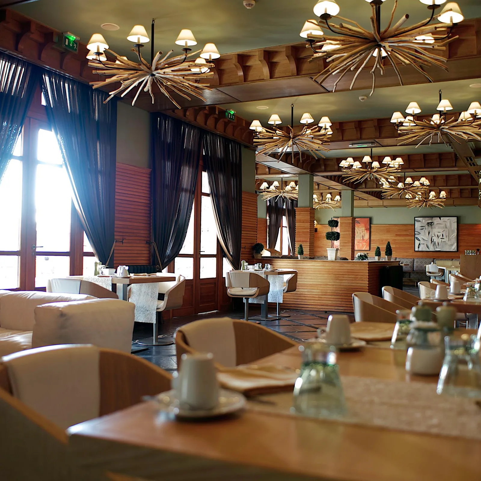 Indoor restaurant with set tables, chandeliers on the ceiling, and large windows draped with dark curtains