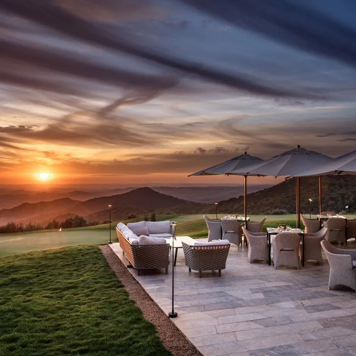Terrace with chairs and tables under umbrellas, facing a sunset over mountains and a golf course.