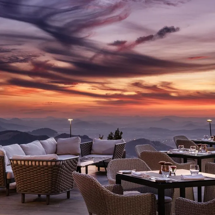 Terrace with wicker tables and chairs, white cushions and glasses, facing a sunset over mountains.