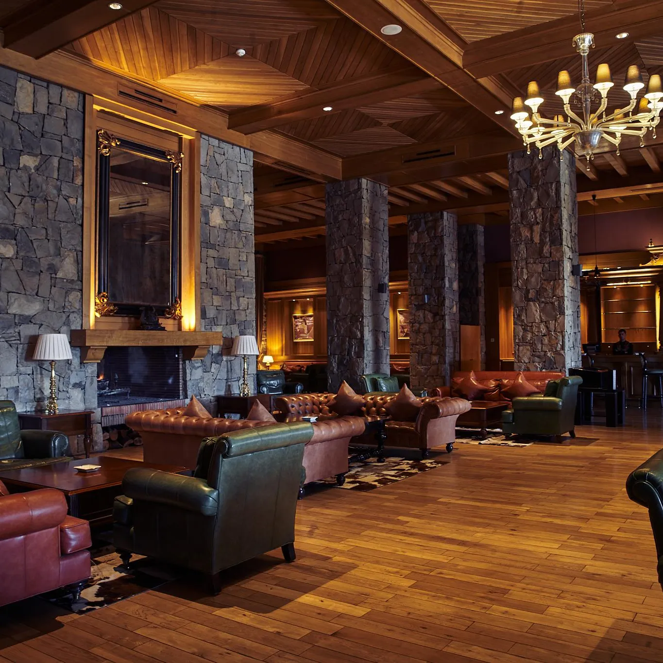 A living room with leather sofas, armchairs, and a stone fireplace, lit by a chandelier and wall lamps.