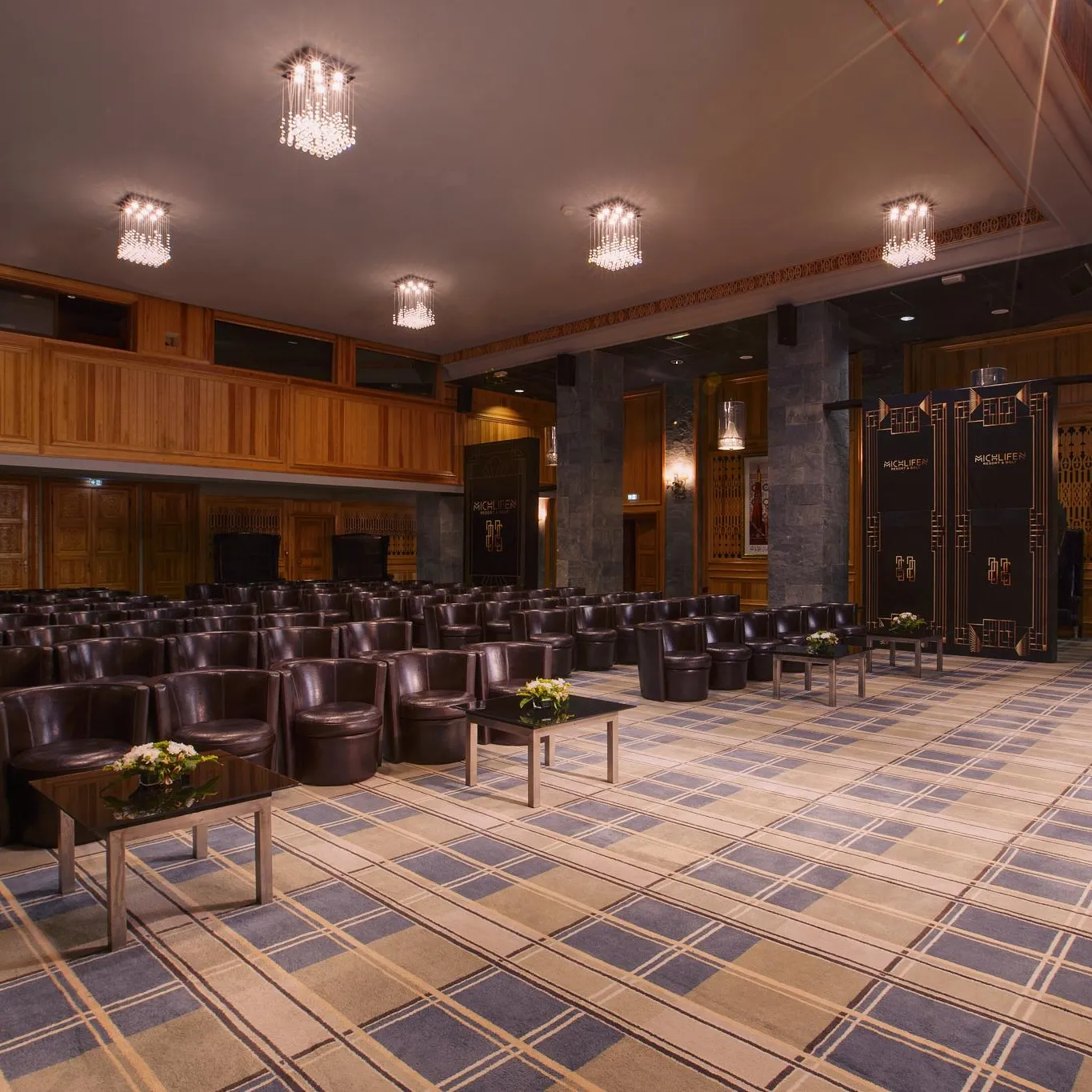Conference room with rows of brown leather armchairs, low tables with flowers, wooden walls, and chandeliers on the ceiling.