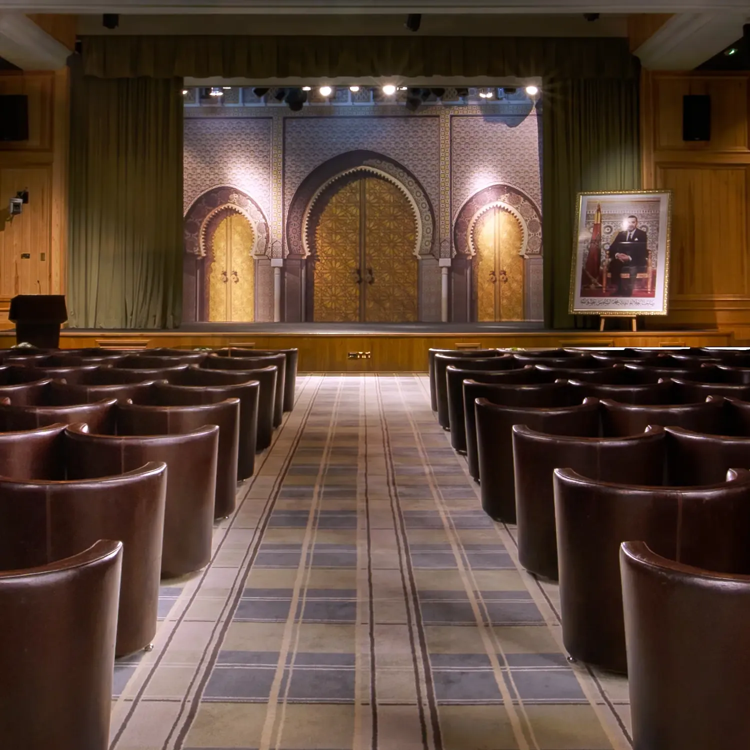 Conference room with leather seats arranged in rows facing a stage adorned with architectural motifs and lit, with a portrait on an easel to the right.