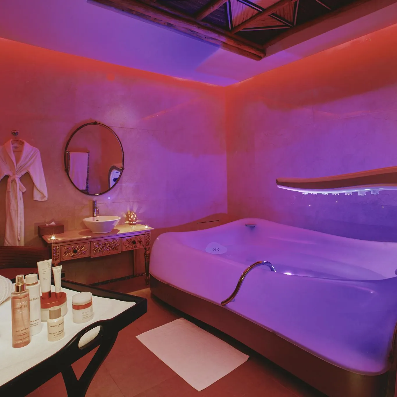 Spa room with a relaxation bathtub, a sink, cosmetic products on a table, and a bathrobe hanging on the wall.