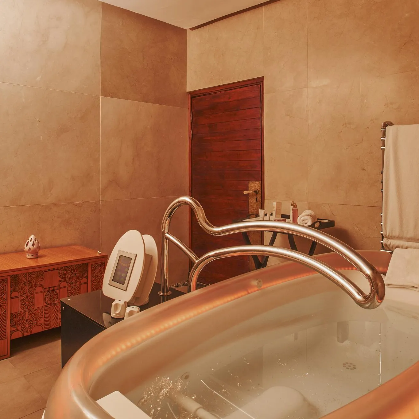 Bathtub filled with water and bubbles, equipped with a faucet and a showerhead, in a bathroom with beige walls, a wooden cabinet, and a towel rack.