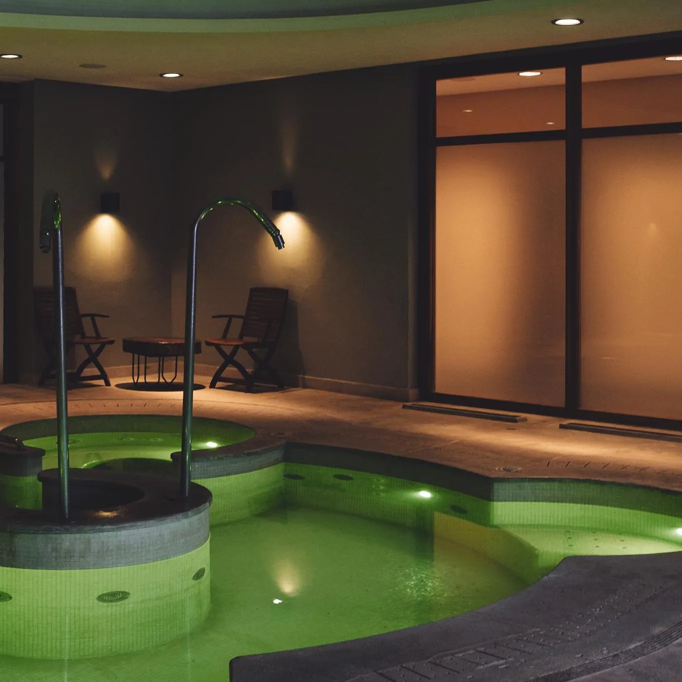 Indoor pool lit in green with two chairs and a table in an enclosed space with dark walls