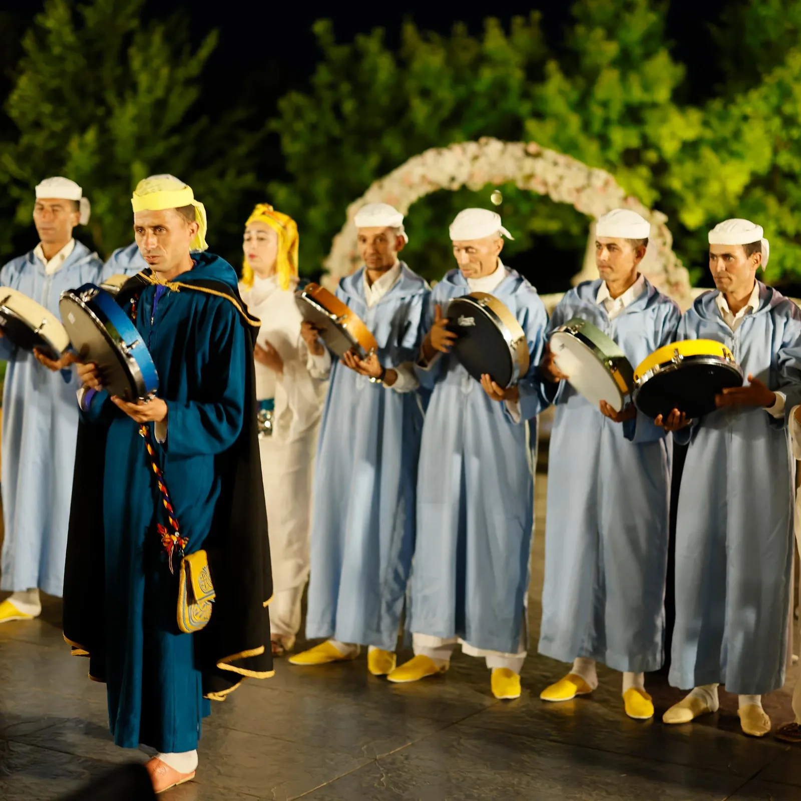 A group of people in traditional costumes plays small drums on an outdoor stage decorated with flowers.