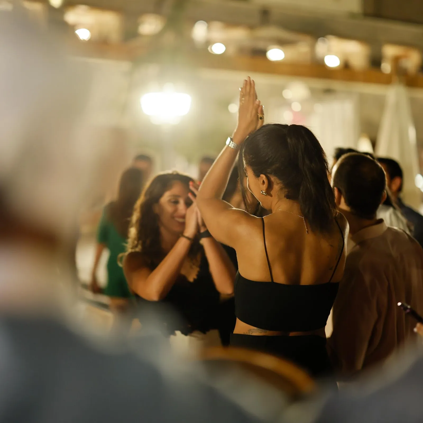 Two women are dancing together in a lit room, surrounded by other people socializing.