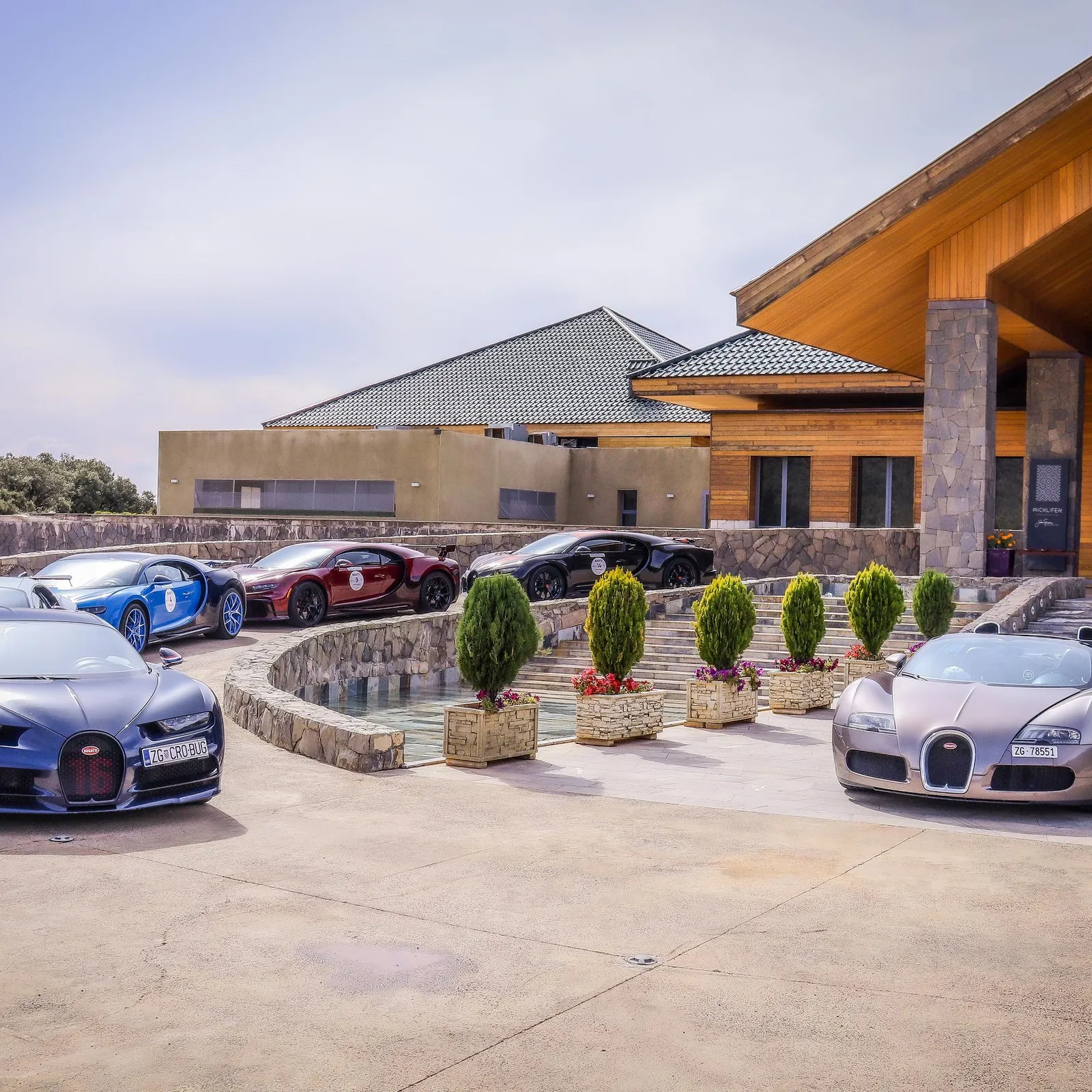 Several luxury cars are parked in front of a stone and wood building with a green-tiled roof.