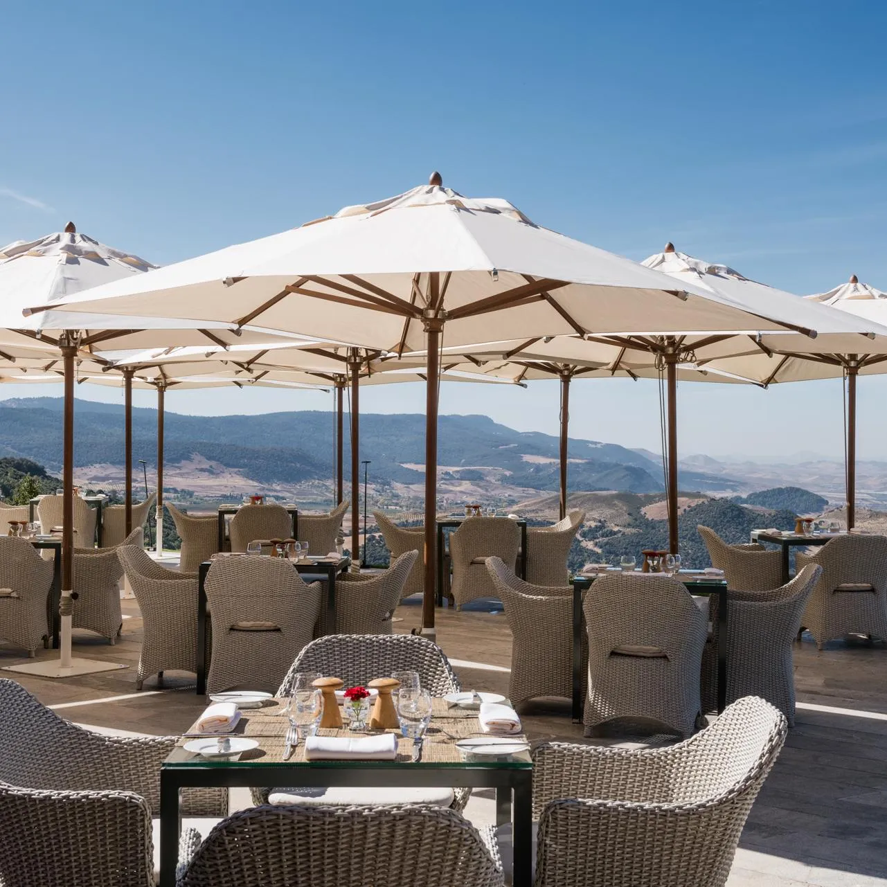 Outdoor restaurant terrace with tables, rattan chairs, and large umbrellas, offering views of mountains and a valley.