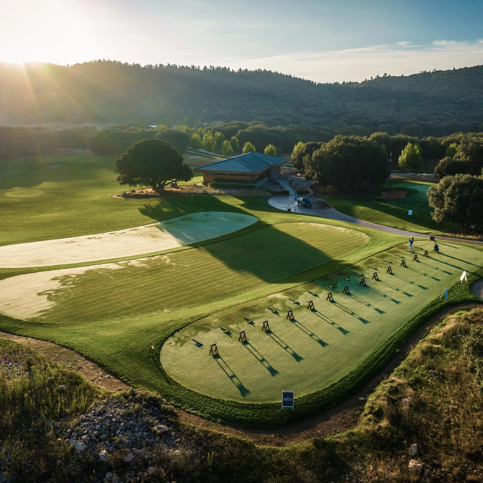 Aerial view of a golf course with players in action, sand bunkers, and a clubhouse in the background under a clear sky.
