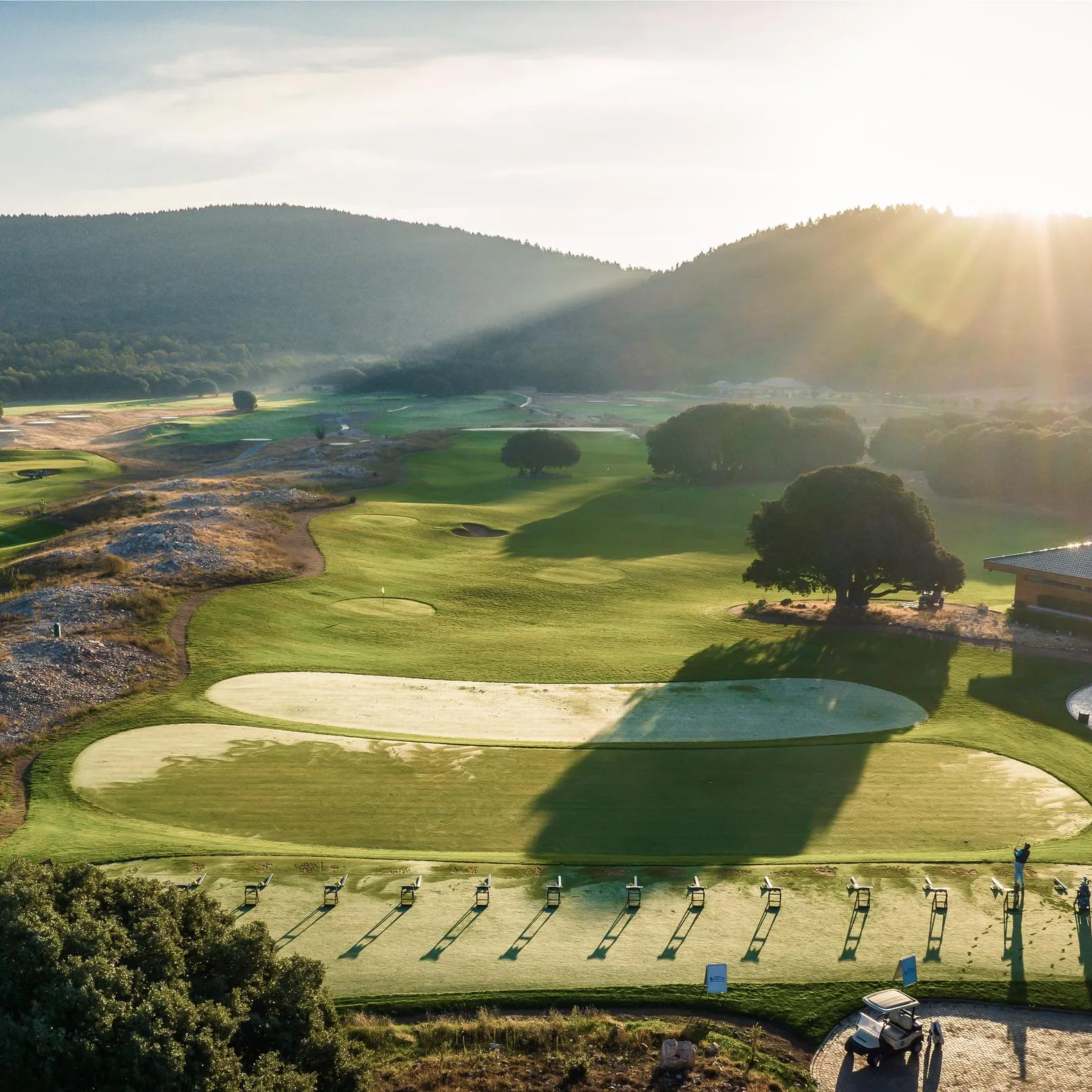A golf course with practice greens and targets, surrounded by wooded hills, under a rising sun