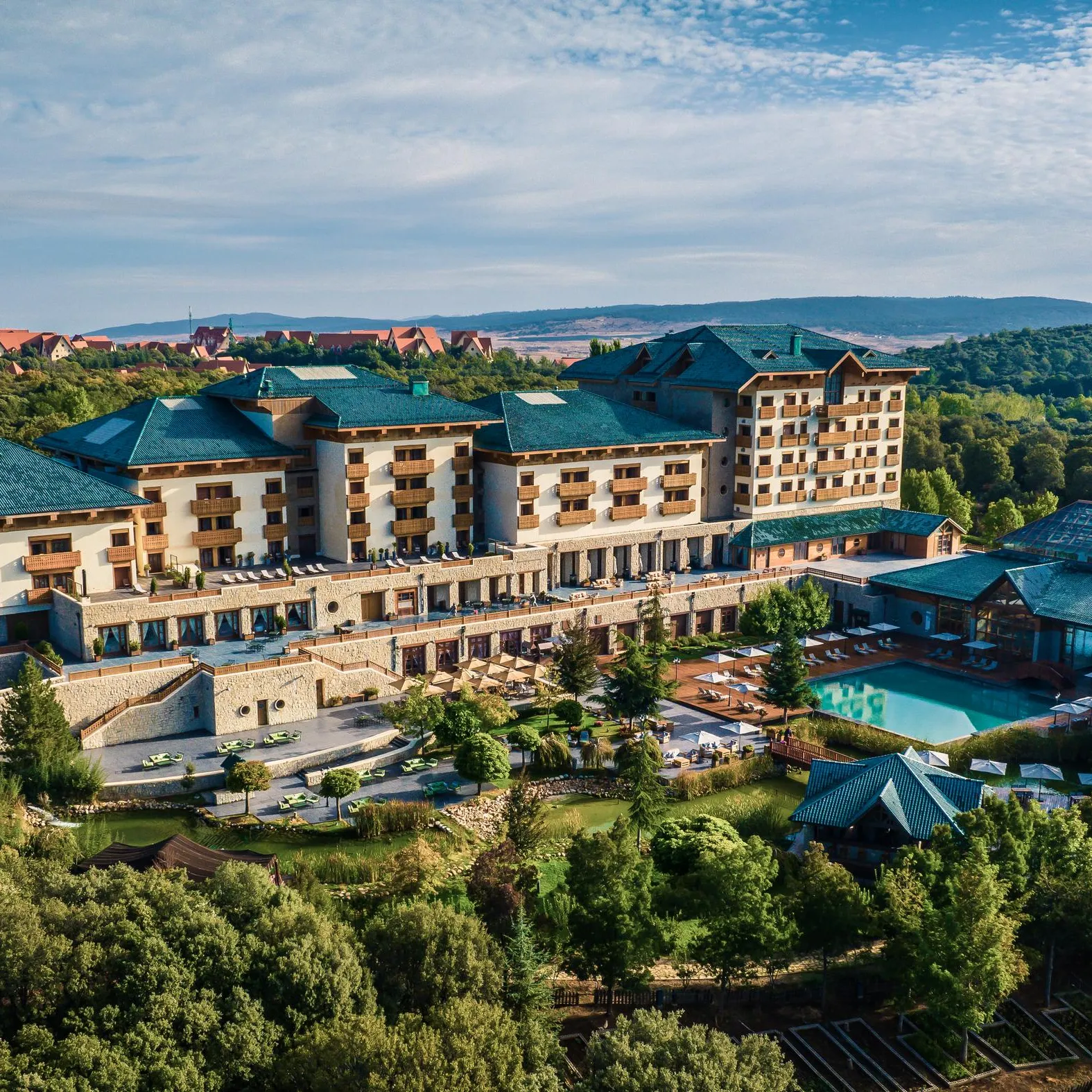 A large hotel complex with green roofs surrounded by trees and gardens, featuring a pool and outdoor relaxation areas.