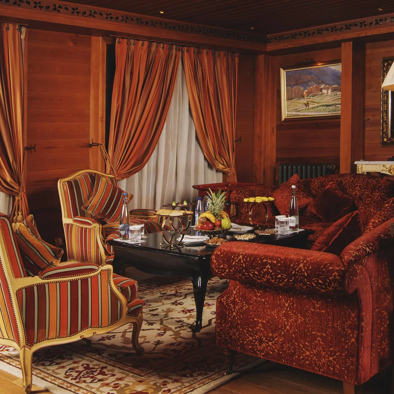 Living room with wooden furniture, striped armchairs, and a red velvet sofa, decorated with cushions and coffee tables featuring fruits and drinks.