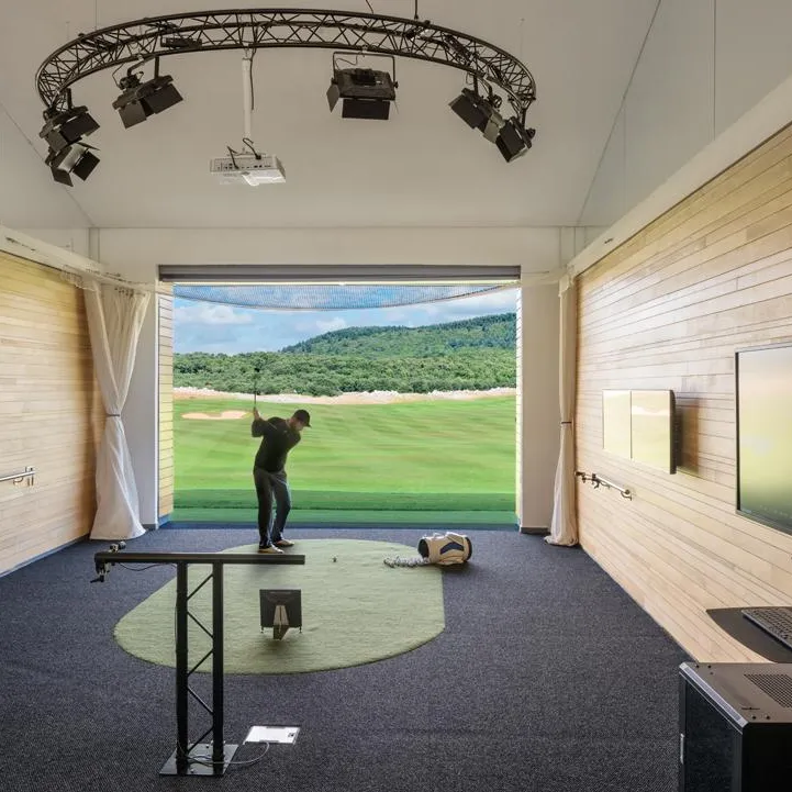 A golfer practices in an indoor golf simulator with a screen projecting a course, equipment, and a monitor nearby.