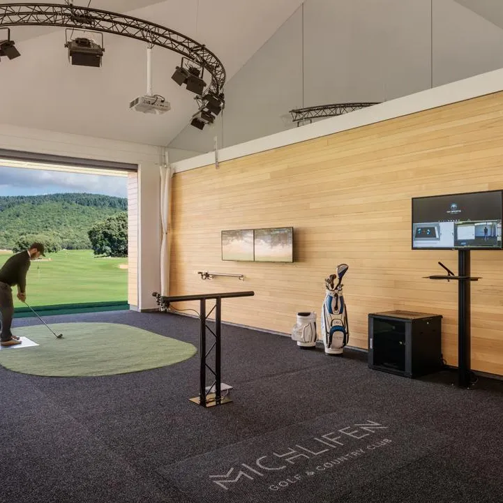 A golfer practices in an indoor training room equipped with a screen projecting a course, a monitor, and a tee mat.