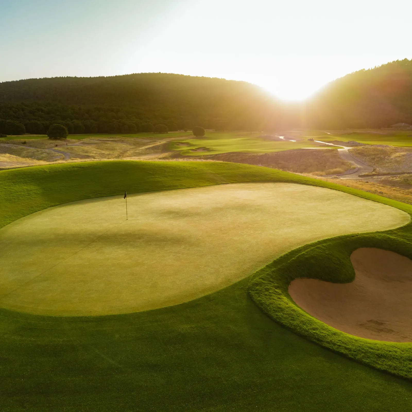 A golf green with a flag in the center, surrounded by sand bunkers, under a setting sun with wooded hills in the background