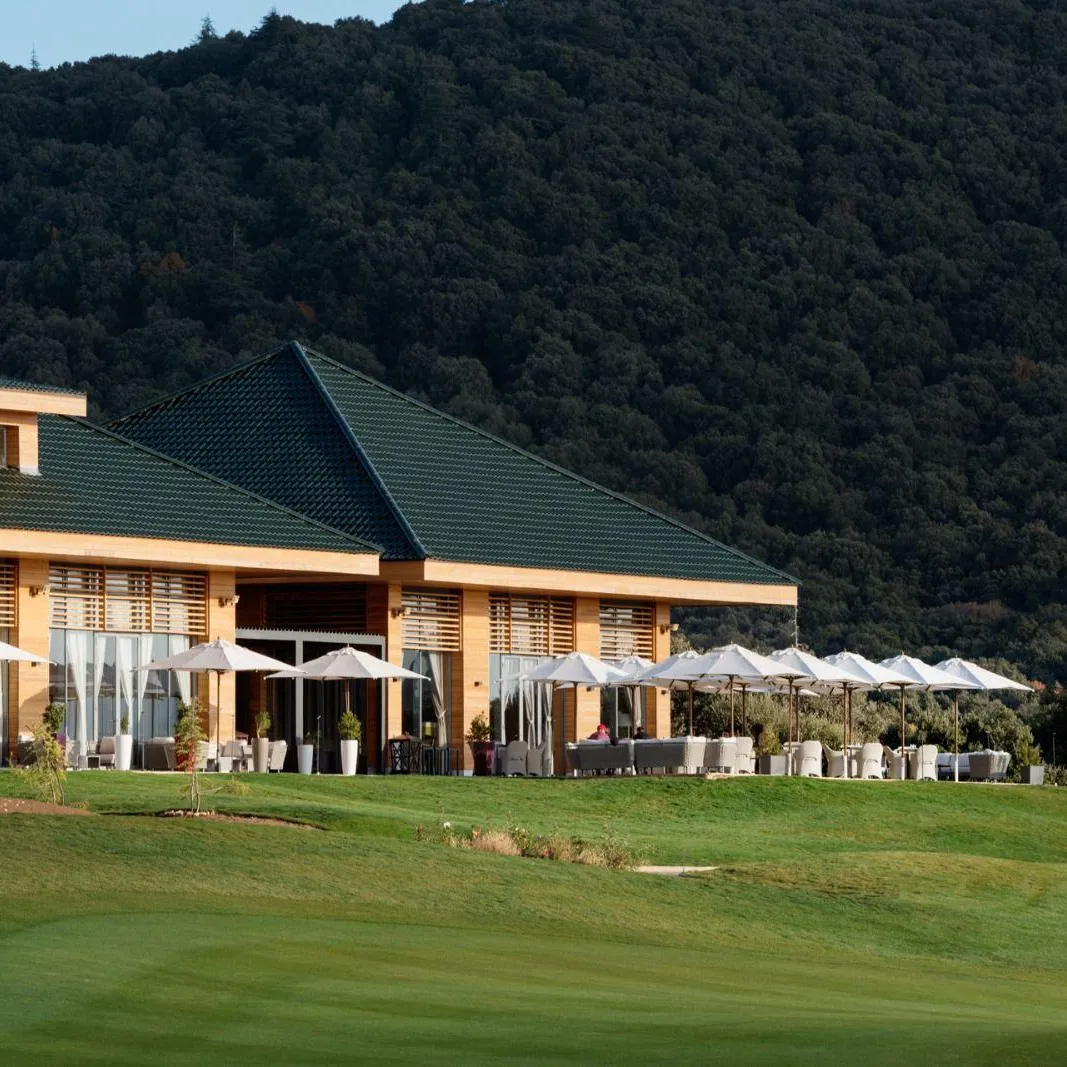 A building with a green roof and outdoor terraces equipped with tables and chairs under umbrellas, located near a golf course with a wooded hill in the background.