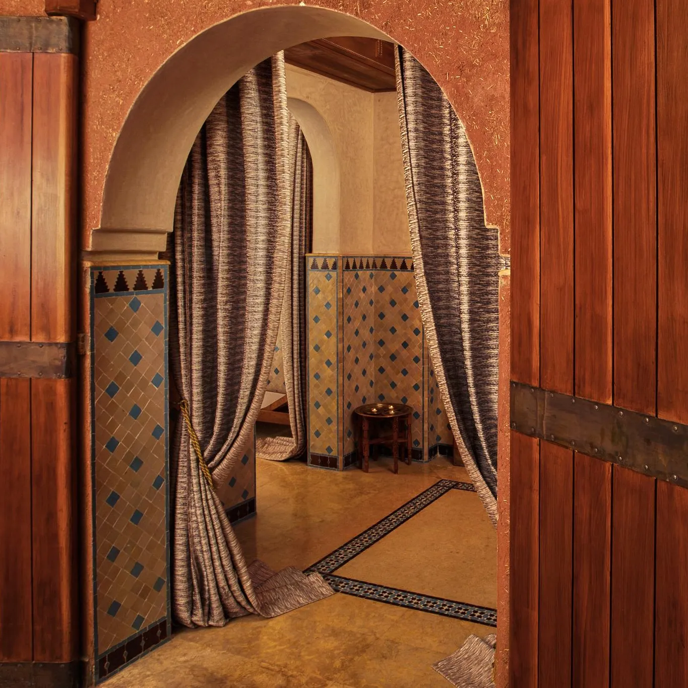Two wooden doors with metal rings open onto a room with tiled walls and arches draped in fabric.