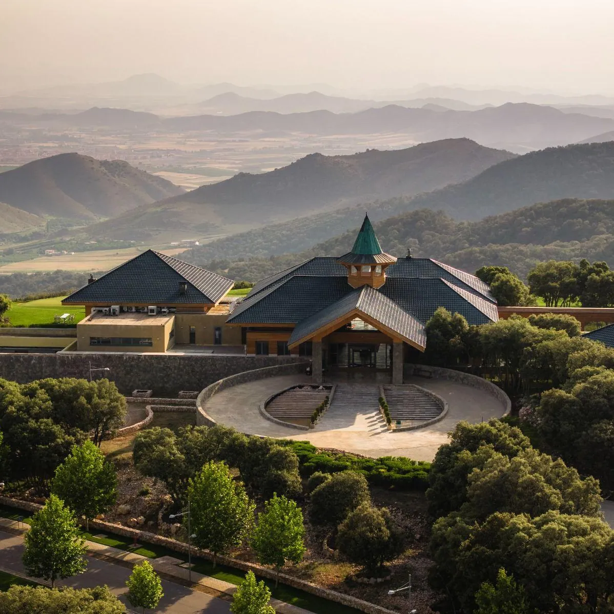 A building with steep roofs and a central tower stands in a rolling, green landscape, surrounded by trees and gardens.
