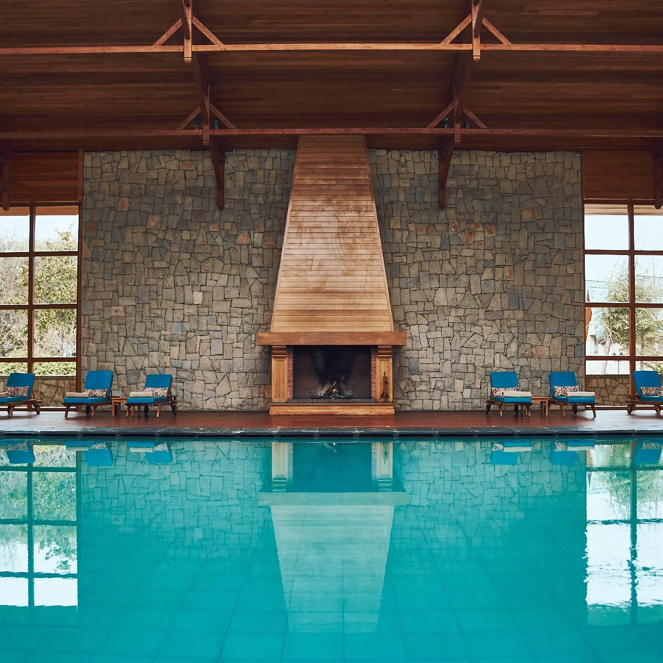 Indoor pool with sun loungers in front of a stone wall and a lit fireplace, surrounded by large wooden windows