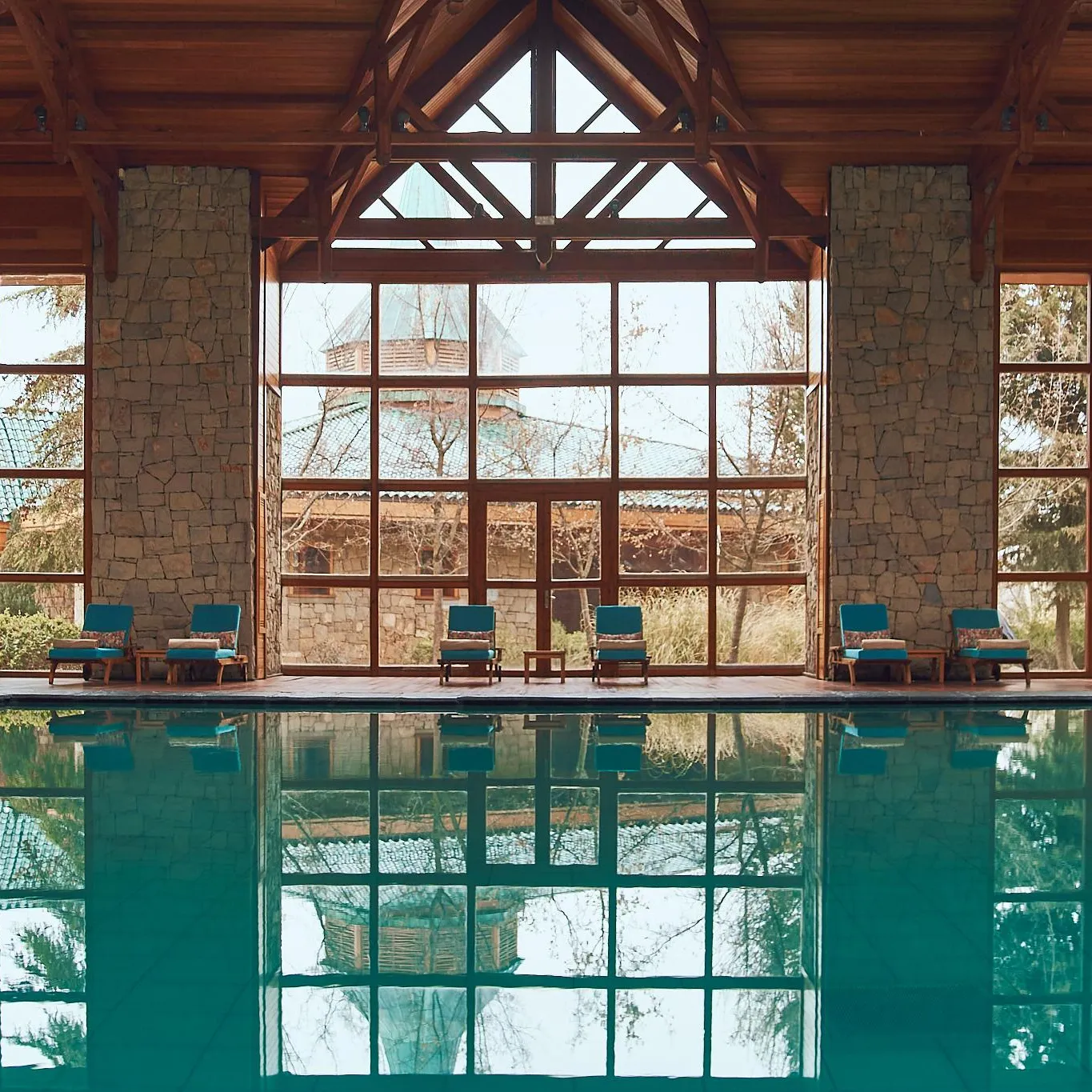 Indoor pool with a view of lounge chairs and stone and wood structures through large windows