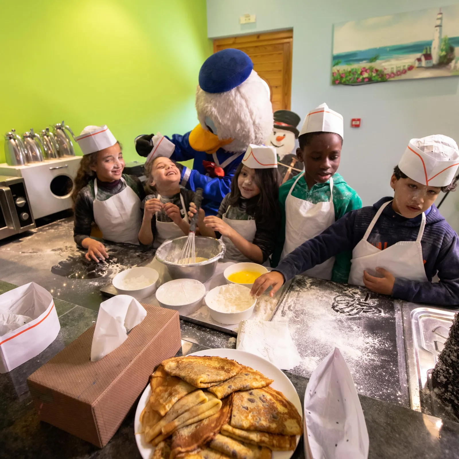 A group of children and a mascot character cook together in a kitchen, surrounded by ingredients and baking utensils.