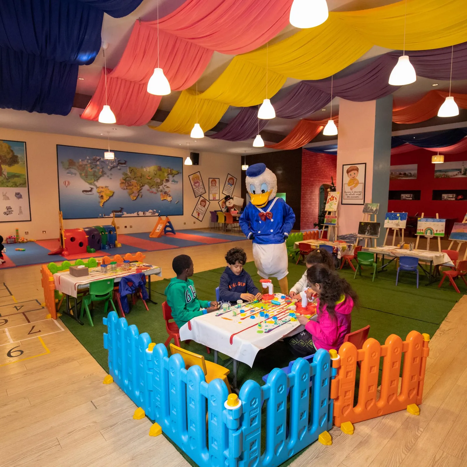 A colorful playroom with children seated at tables, a cartoon character in costume, and educational wall decorations.