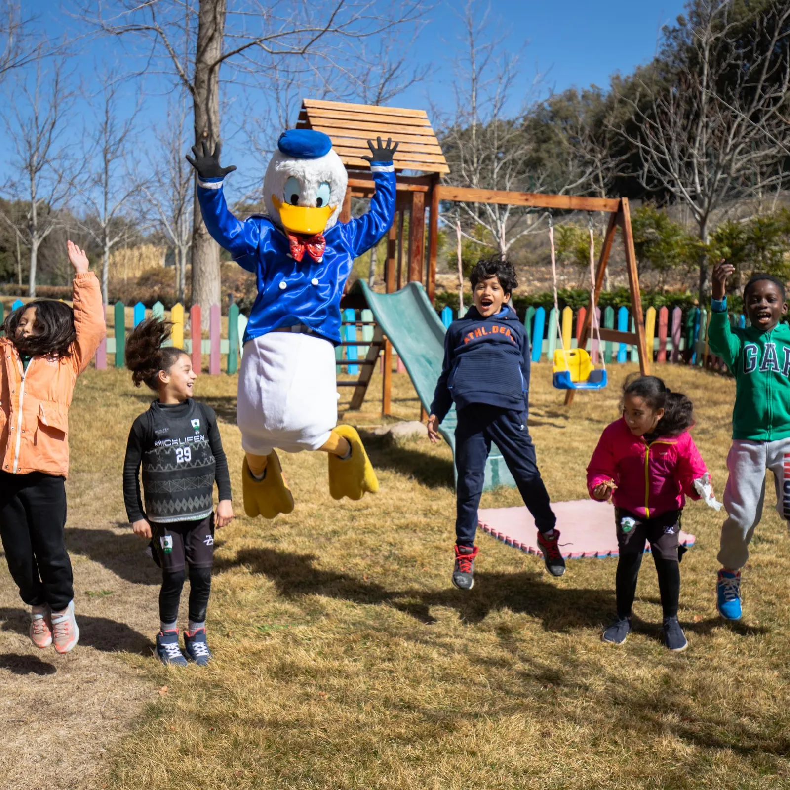 A costumed duck character jumps with five children in a park with wooden games and a colorful fence in the background.