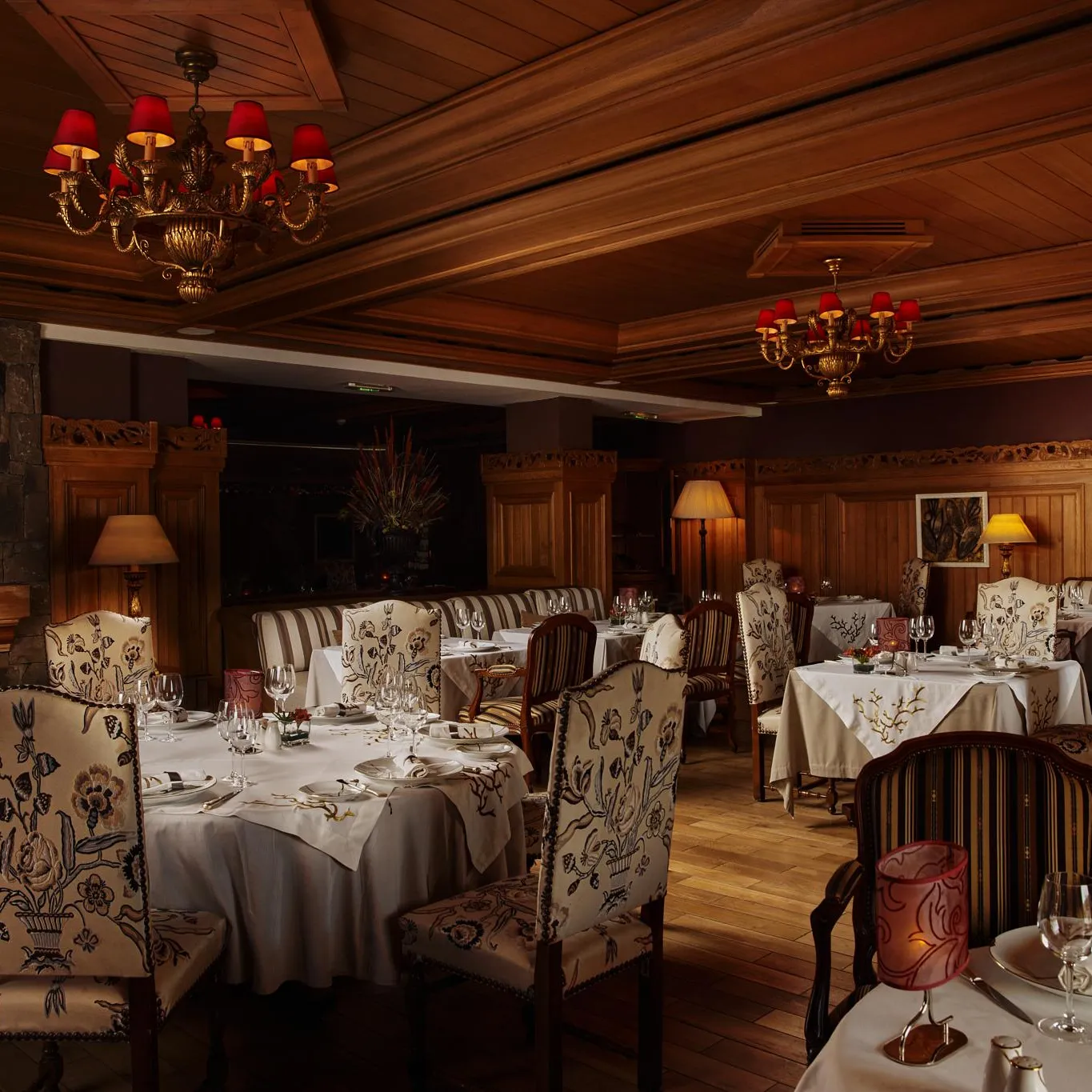 Elegant restaurant with set tables, chandeliers, and floral-patterned chairs, lit by lamps and chandeliers
