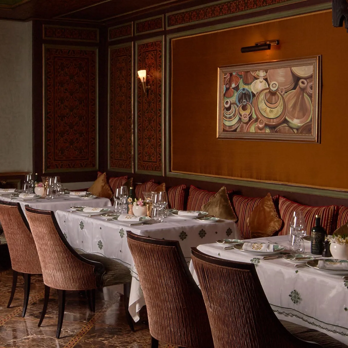 An elegant dining room with set tables and upholstered chairs, decorated with cushions and a colorful wall painting.