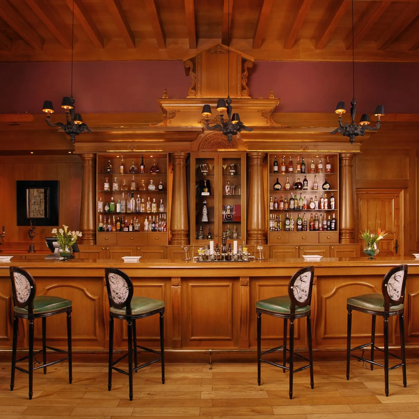 A wooden bar with shelves filled with bottles, green stools, and flowers on the counter.