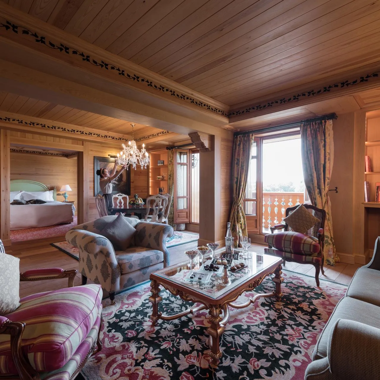 Luxurious wooden room with a seating area, a coffee table, padded chairs, and a bed visible in the background.