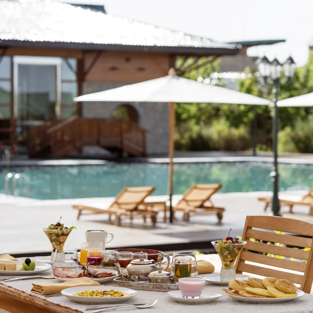 Breakfast table on a terrace with food, drinks, and fresh fruit, overlooking a pool with sun loungers and umbrellas