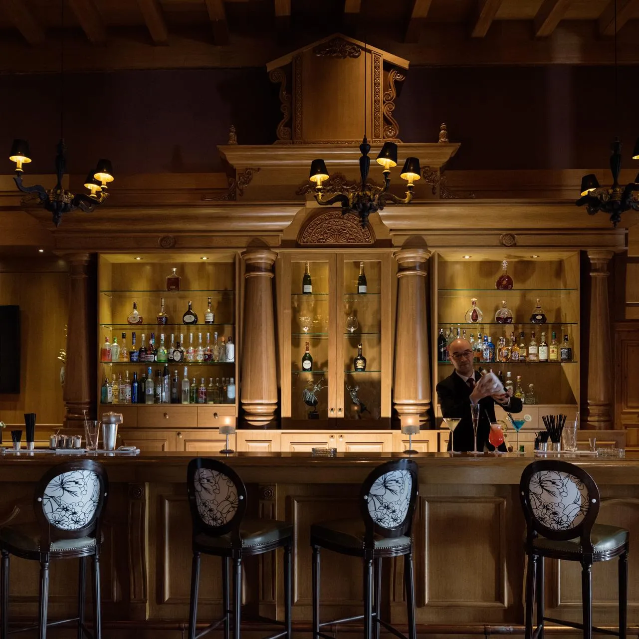 A bartender prepares a cocktail behind a wooden counter with shelves filled with bottles and glasses.