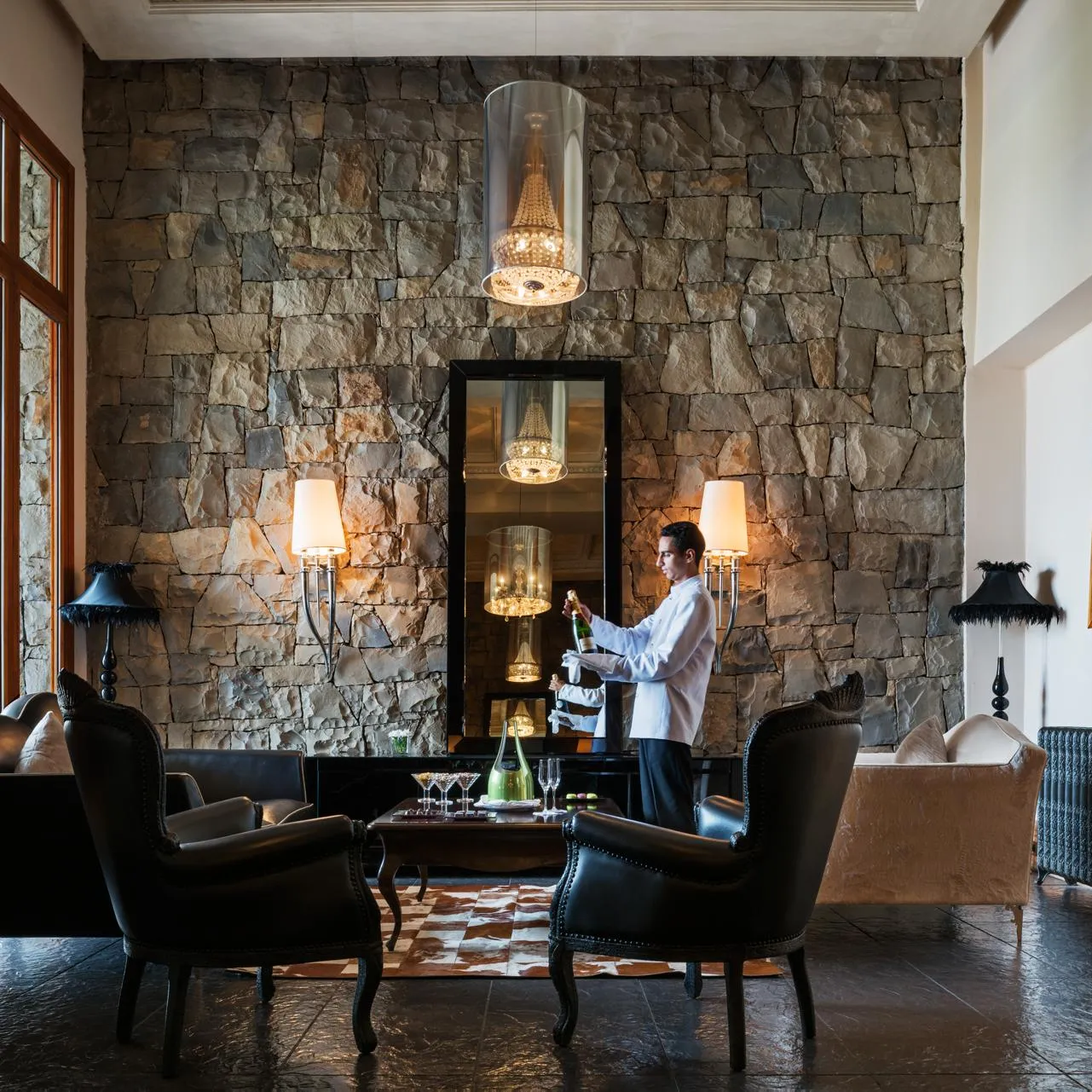 A waiter in a white jacket prepares drinks behind a stone bar with candelabras, surrounded by armchairs and lamps in an elegant lounge.