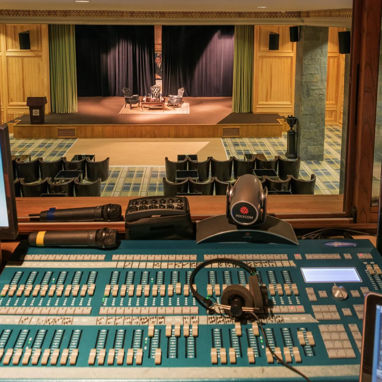 Sound mixing desk with screens and microphones in front of a theater stage with chairs and a closed curtain