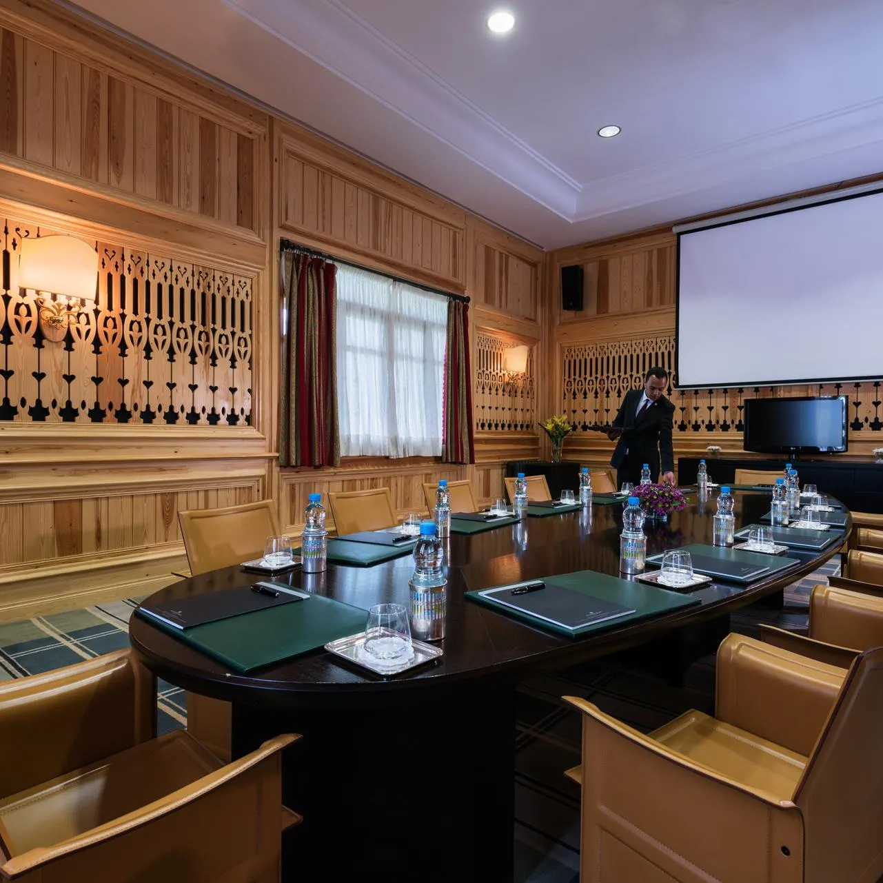 Wooden meeting room with an oval table, leather chairs, a projector, and a man standing near a screen