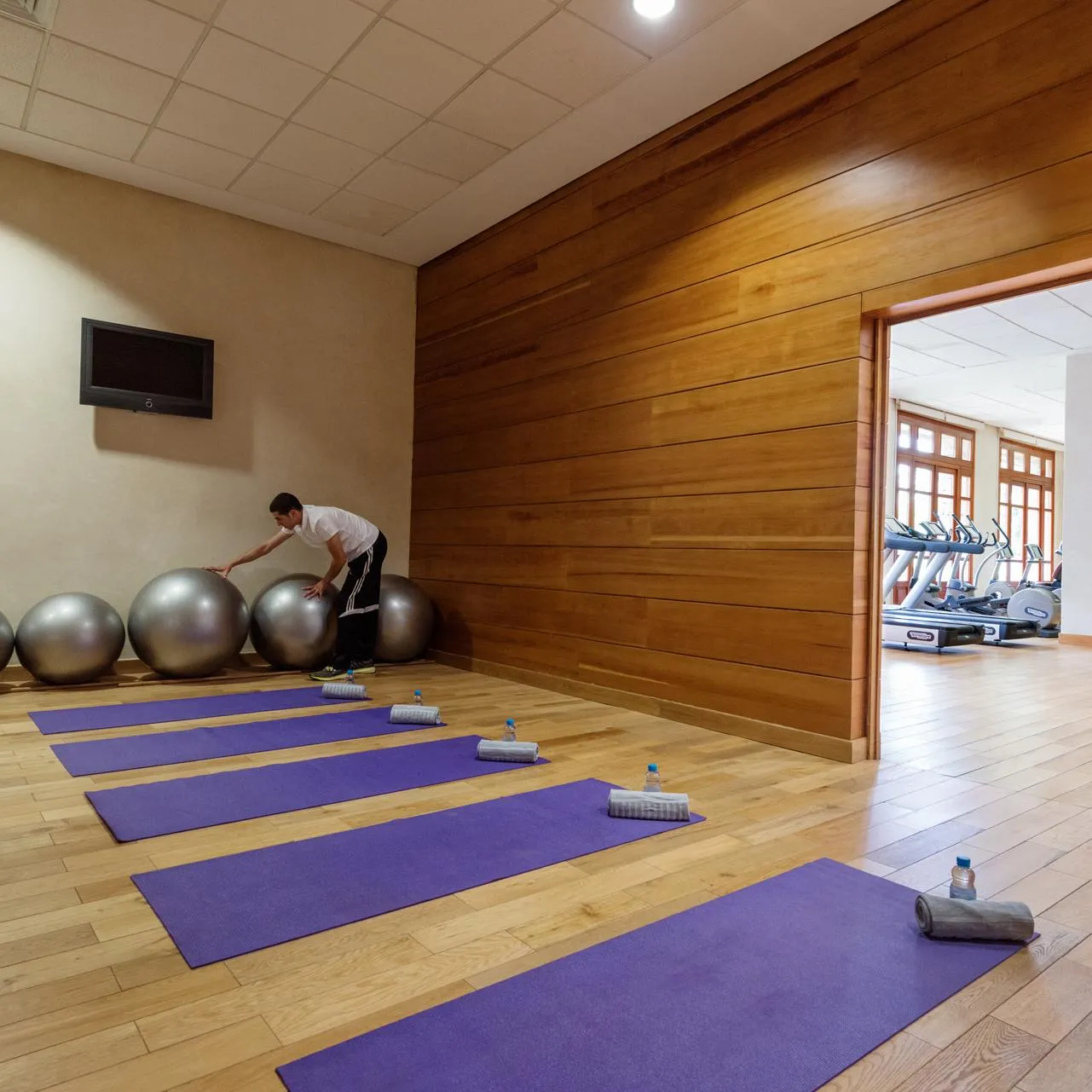 A man uses an exercise ball in a gym with yoga mats on the floor and fitness equipment visible through an open door.