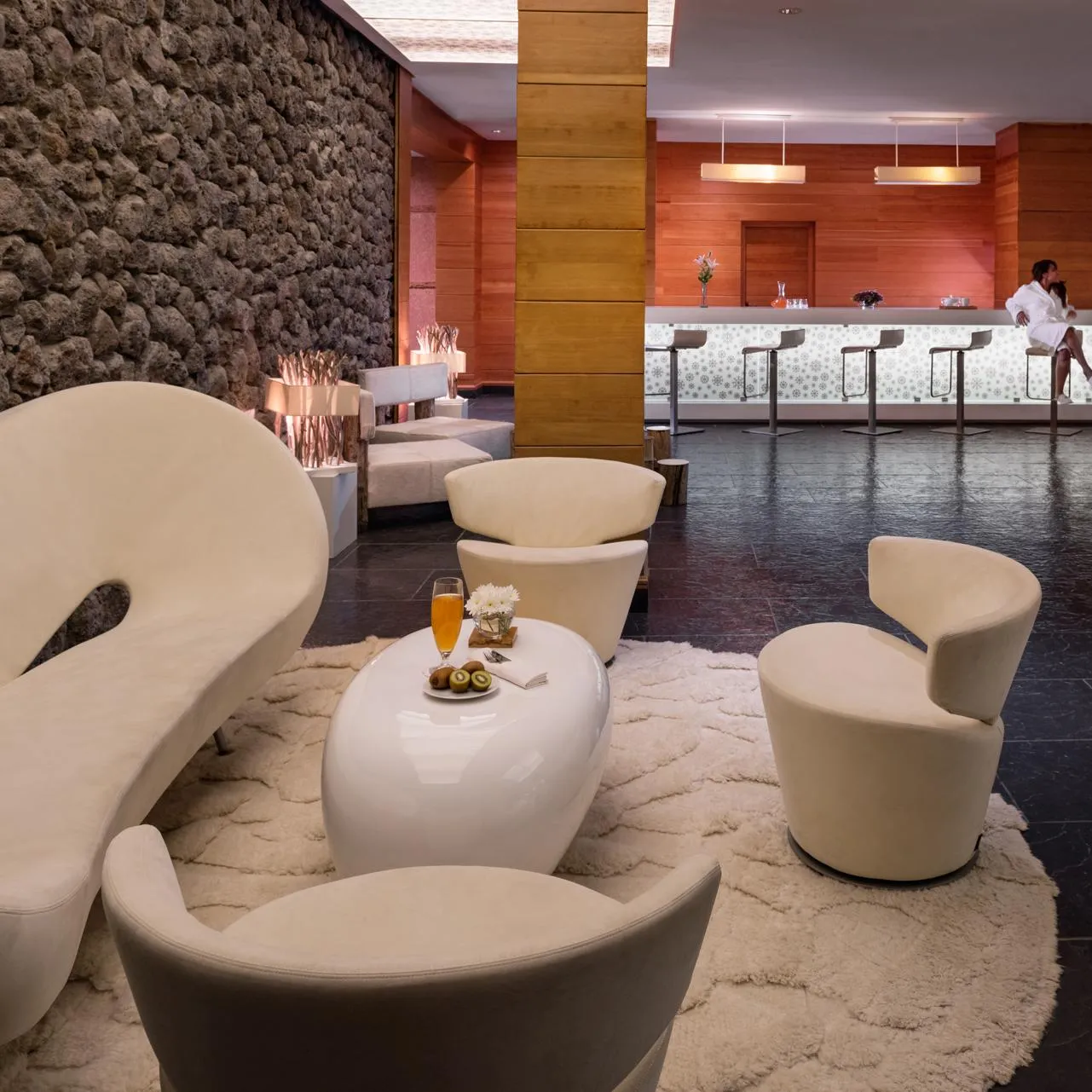 A hotel lounge with modern seating, a coffee table with drinks and fruit, and a bar with stools in the background.