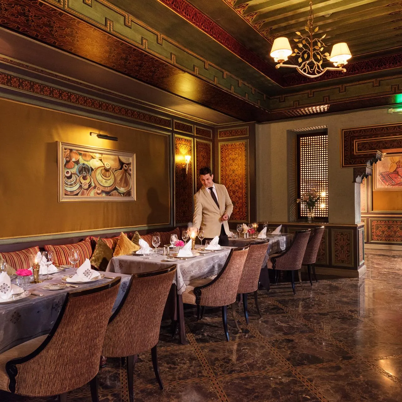 A server in a beige suit bustles in an elegant restaurant with walls adorned with patterns and paintings, with tables set for service.