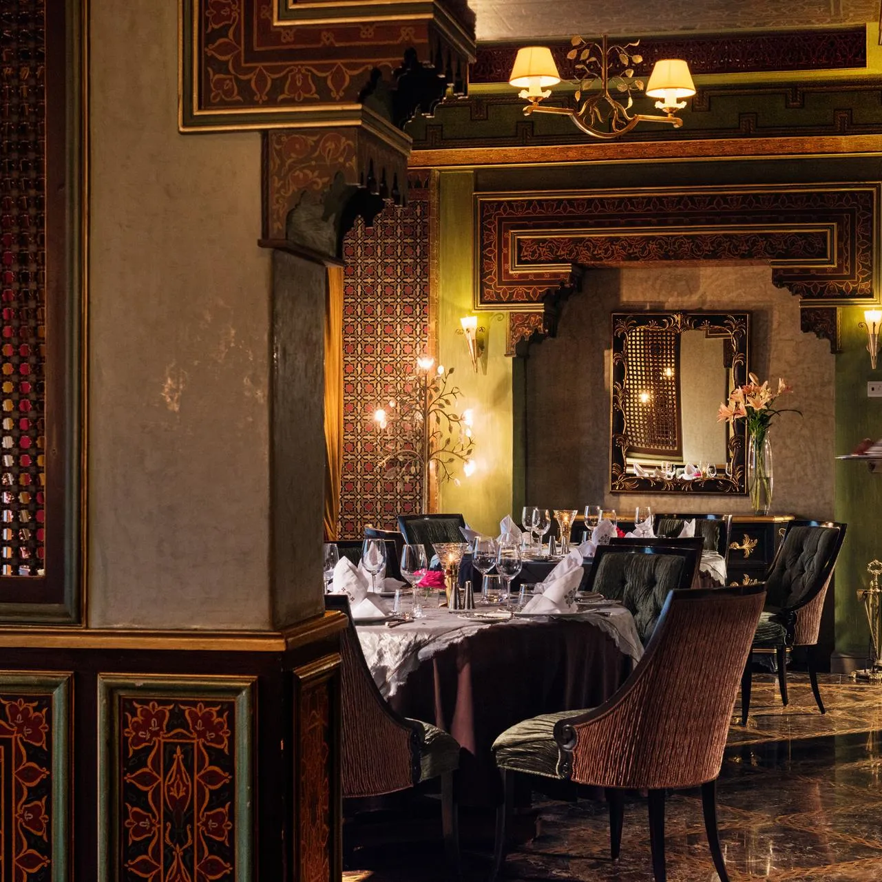 A waiter in a suit carries a tray in an elegant restaurant with walls decorated with patterns and lit by wall lamps.