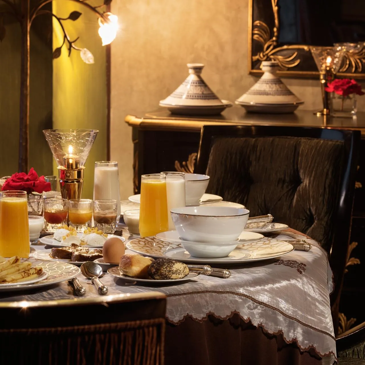 Table set for breakfast with drinks, eggs, pastries, and fruit, in an elegant setting with candelabras and cushions.
