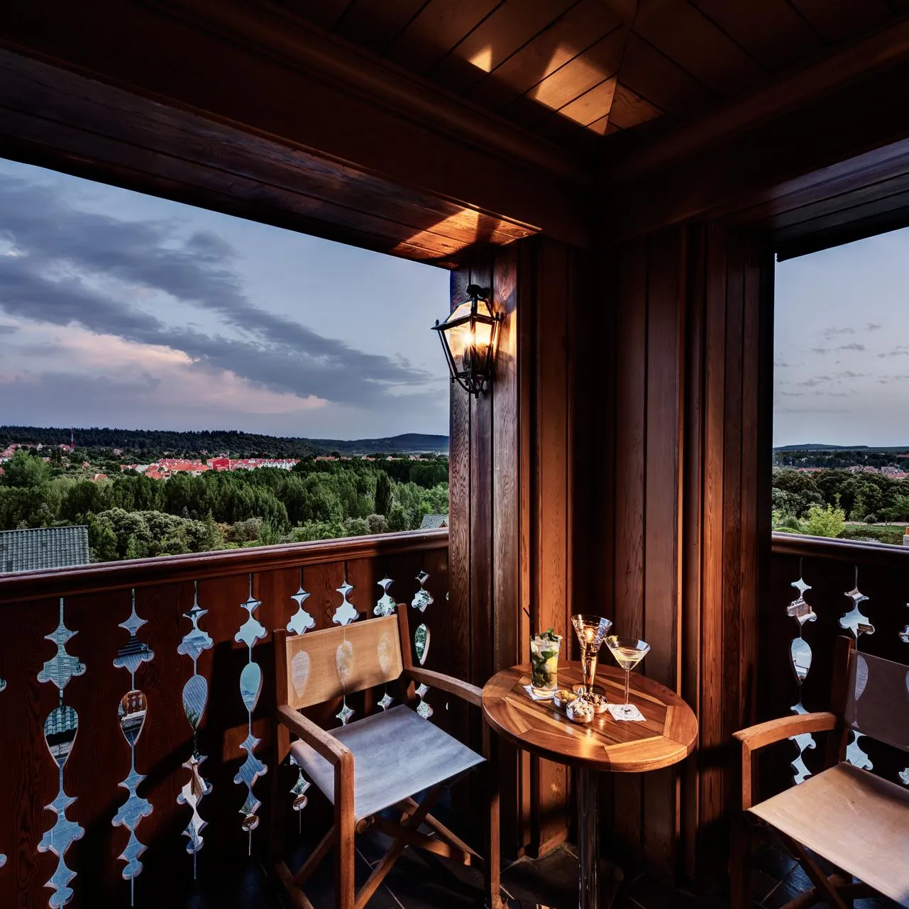 Wooden balcony with table, chairs, and glasses, offering a panoramic view of a city and hills under a cloudy sky.