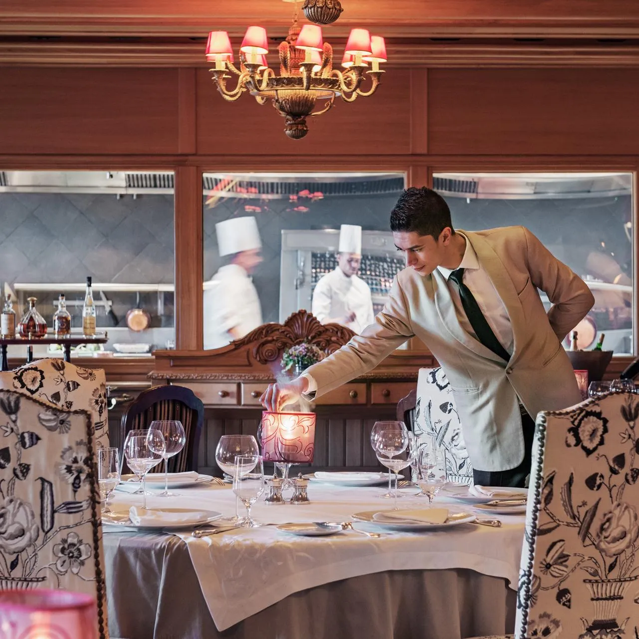 A waiter in a suit lights a candle on a table set in an elegant restaurant with an open kitchen in the background.