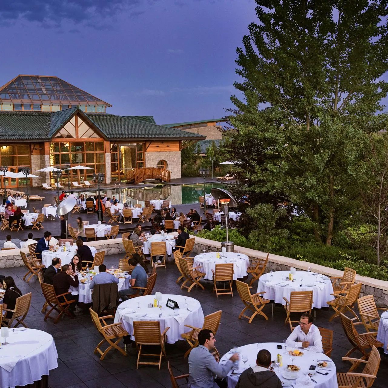 An outdoor restaurant terrace with many tables and chairs, diners eating, and a wooden and stone building in the background.