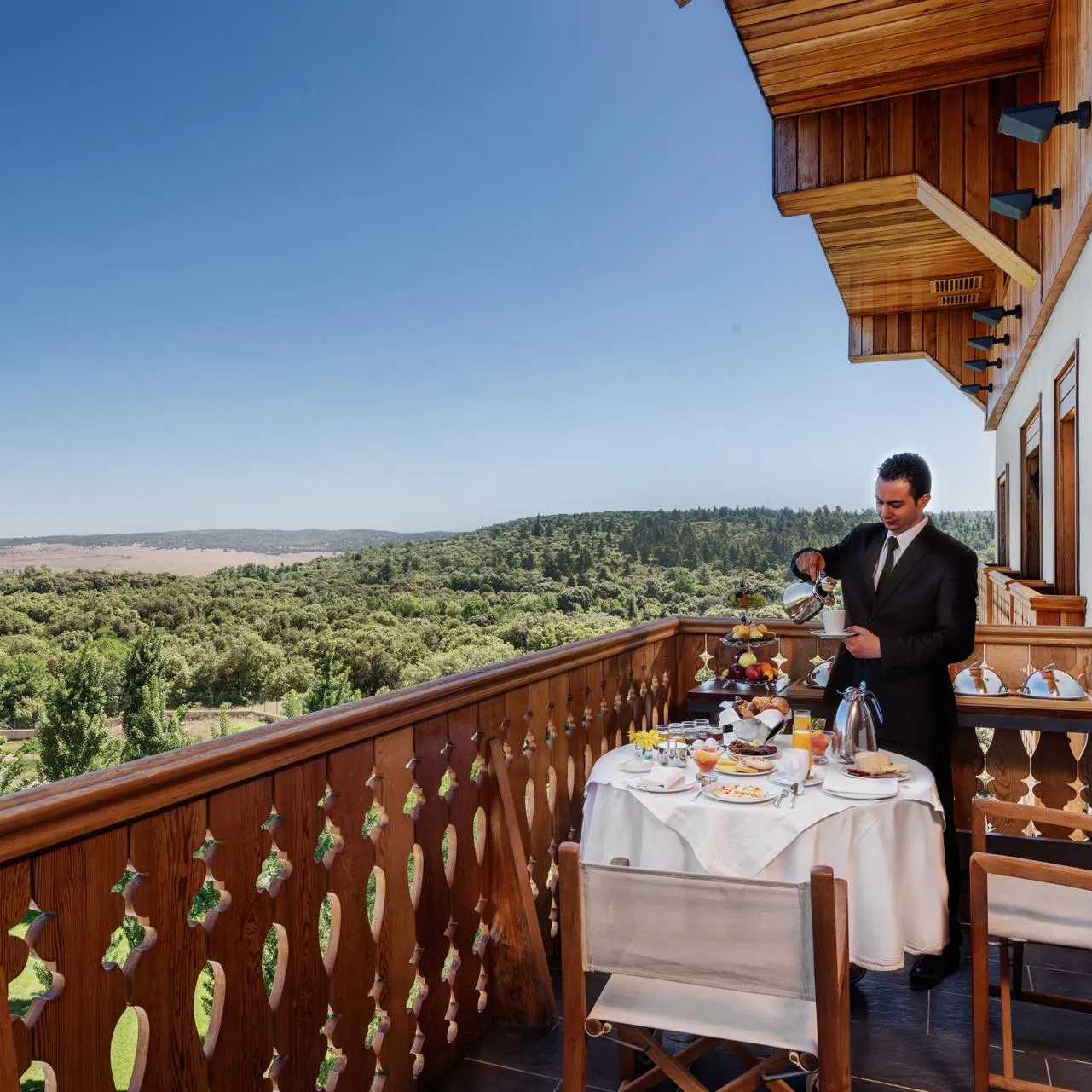 A waiter in a black suit serves a meal on a wooden terrace with a view of a forest and hills.