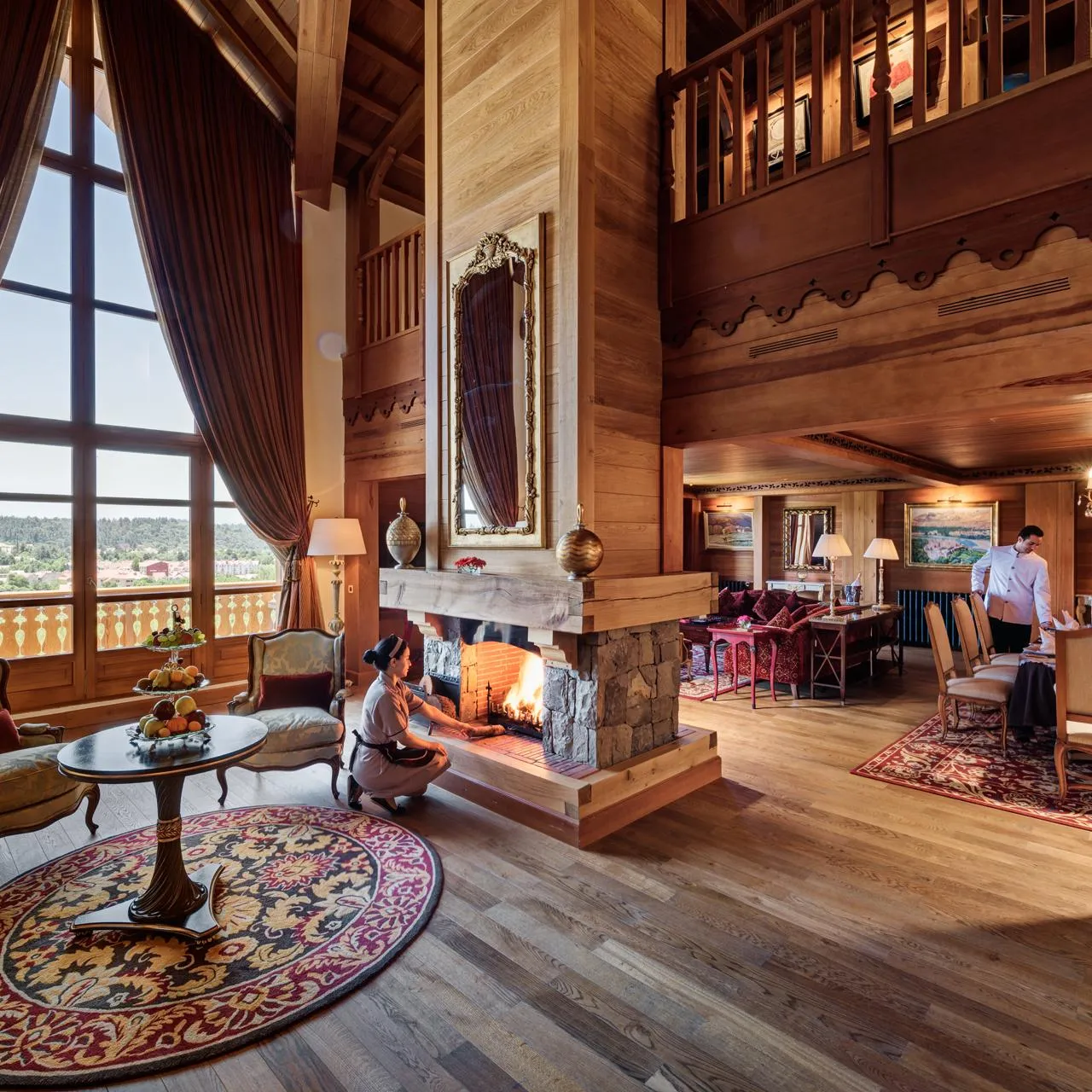 A wooden living room with a large window, a lit fireplace, armchairs, a coffee table, and people near the fire and the dining table.