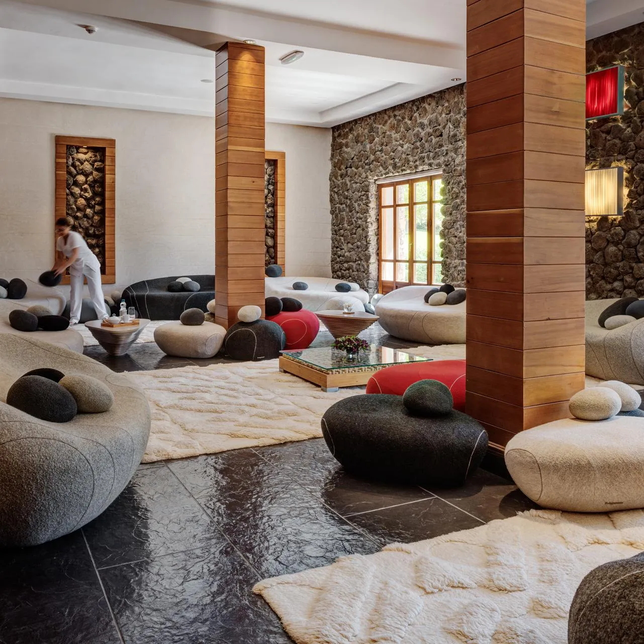 A person in white clothing is cleaning the floor of a room with giant cushions and stone and wood walls.