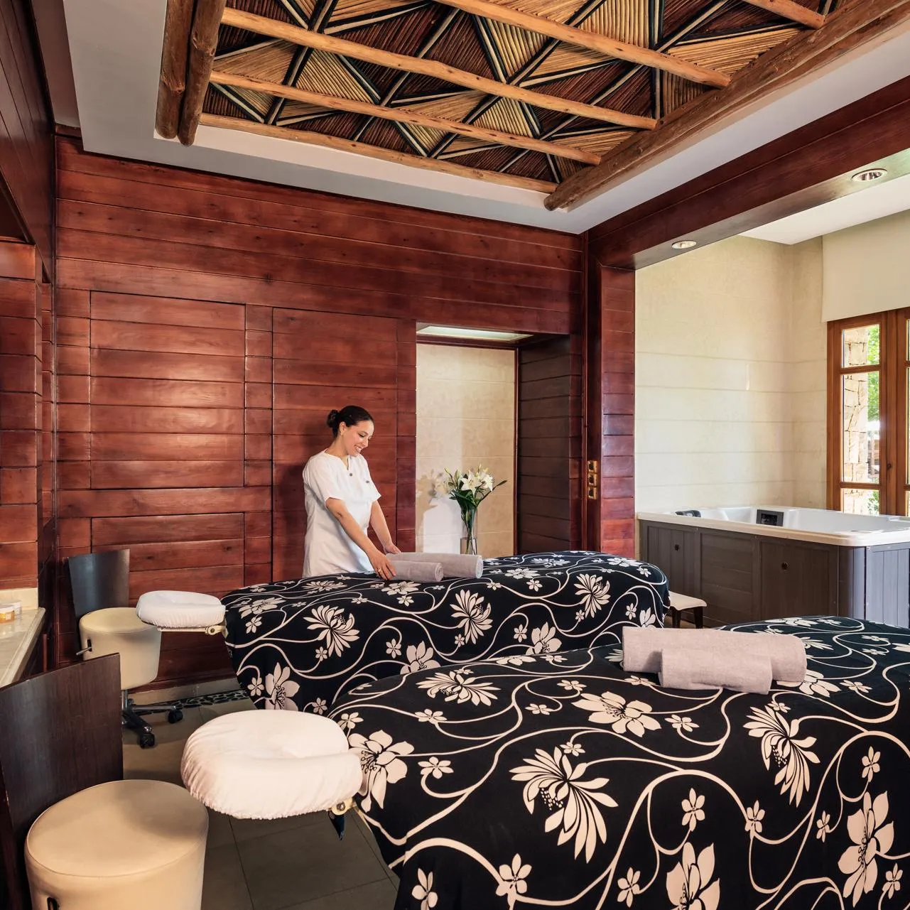 A person in white attire prepares a massage table in a wooden spa room with windows and plants.