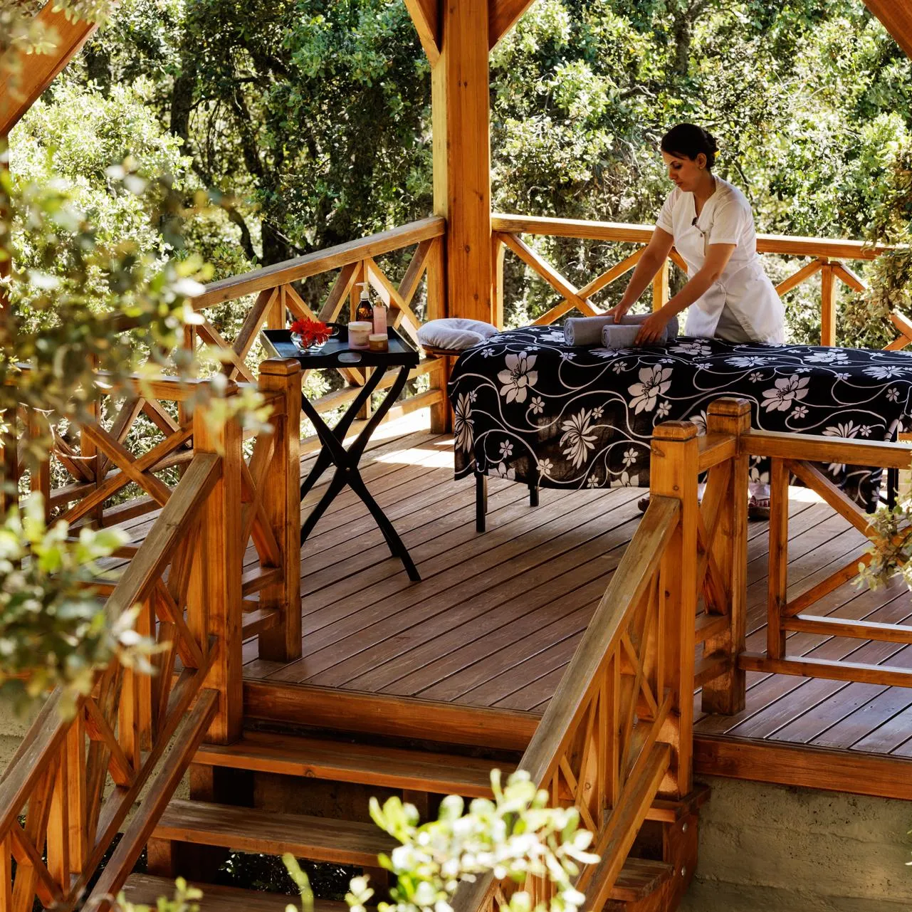 A person in white clothing is giving a massage to a person lying on a table outside, surrounded by trees.